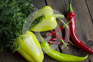 On a wooden table are two bell peppers, hot green peppers, hot red peppers and many small chillies with dill. Different types of