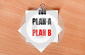 On a wooden table under a black paper clip lies a sheet of white paper with the text PLAN B