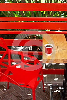 A wooden table on which there is a red plastic cup with coffee. Summer beautiful terrace in greenery. Cozy place