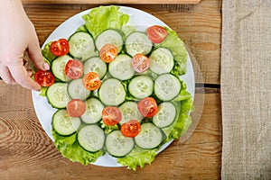 On a wooden table, a woman puts the cherry tomatoes on a plate of cucumbers.