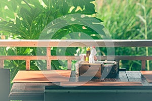 Wooden tissue box and garnish in basket salt, pepper, toothpick, sauce on table with green trees and sunlight background.