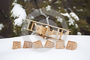 A wooden toy airplane in the snow, against the background of a forest and the word Russia made up of cubes. The concept