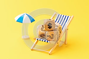 Wooden toy cubes with dollar symbol and question mark in deck chair and parasol on yellow background, vacation cost concept