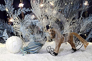 Wooden toy rocking horse stands on artificial snow and New Year toys lie next to it and artificial blue and white trees