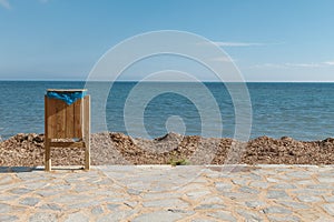 Wooden trash can with a bag on the boulevard of a public city beach on the background of garbage and clear blue sea and sky