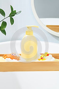 A wooden tray is placed across the bathtub, above it displays a bottle of shower gel, a plastic crab and two silicone ducks, a