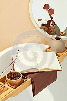 A wooden tray rests across a bathtub, adorned with a wooden bowl of coffee beans, an open notebook, and a vase of flowers.