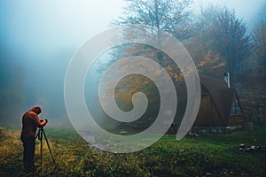 Wooden triangle hut in the mountains forest. Autumn colors. Dense mist fog. Man photographs the landscape on a tripod.