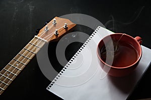 Wooden ukelele on a black desk next to a mug with a hot drink and a notebook for inspiration concept on a music production studio.