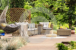 Wooden veranda with rattan furniture, comfy swing and trees in t