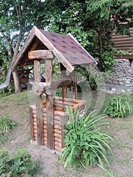 A wooden village well, made of cylindrical logs with a flexible tile roof, a wooden wheel and old rusty chain. Country style