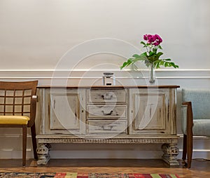 Wooden vintage off white sideboard, glass vase with red flowers, two chairs on white wall and wooden parquet floor