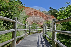 Wooden walkway in the midst of vegetation, in the background an orange cliff. Vibrant blue sky without clouds.