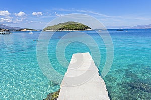 wooden walkway over the crystal clear waters with an islet in the background of the paradisiacal beach of Ksamil in Albania.