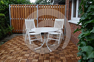 Wooden wall, wooden tiles, two white garden chairs and a round table on  terrace of a residential house