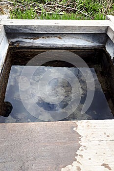 Wooden well with clear water in countryside. Reflection of the sky with clouds in calm water. Vertical photo