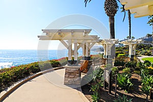 A wooden white pergola surrounded by lush green palm trees and plants with red flowers and lush green grass