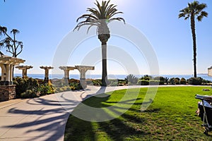 A wooden white pergola surrounded by lush green palm trees and plants with red flowers and lush green grass near blue ocean water