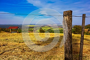 Wooden and wire fence on top of a hill with the landscape of pastures and plantations in the background in sunny day and blue sky