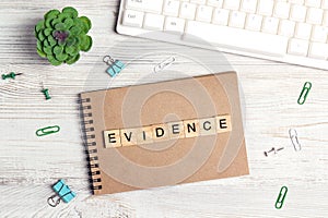 Wooden word Evidence in workspace on paper notepad with computer keyboard, supplies  and green plant on light wood background