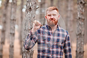 A woodsman stands in the woods among the pines and holds an ax on his right shoulder. Outdoors.