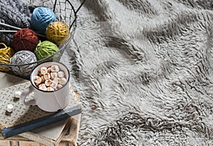 Wool yarn in vintage metal basket, old books and hot chocolate in a ceramic mug on light tissue surface.