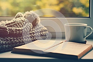 Woolen scarf, a cup of tea and book on the windowsill. Hygge and cozy autumn concept