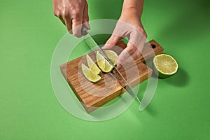 Slices of natural organic green lime, cutting by female hands on a wooden board on green background with copy space.