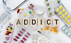 The word ADDICT on wooden blocks on the table. Medical concept with pills, vitamins, stethoscope and syringe in the background