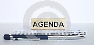 The word Agenda on a bar of jenga wood, lying on a Notepad with a metal blue pen