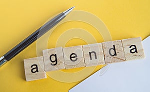 The word agenda is written on wooden cubes between a pen and a white sheet of paper on a yellow background  top view  flat layout