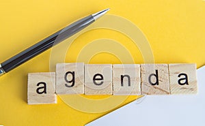 The word agenda is written on wooden cubes between a pen and a white sheet of paper on a yellow background  top view  flat layout