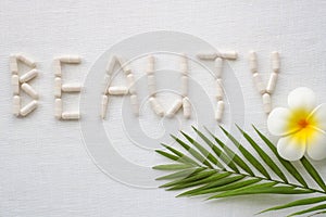 Word BEAUTY made from collagen pills, green palm leaf and plumeria flower on white textile background. Healthy lifestyle