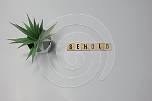 The word Behold written in wooden letter tiles on a white background