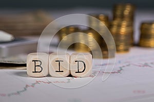 Word BID composed of wooden letter. Stacks of coins in the background