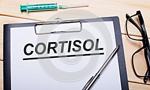 The word CORTISOL is written on a white piece of paper next to black-rimmed glasses, a pen and a syringe. Medical concept