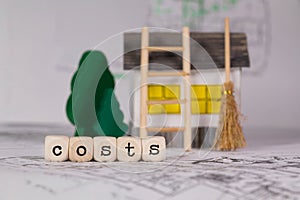 Word COSTS composed of wooden letter. Small paper house, wooden trees in the background