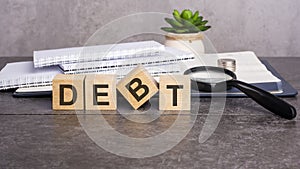 the word Debt is written on wooden cubes on a gray background. close-up of wooden elements, magnifying glass, paper