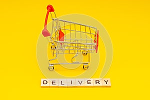 Word DELIVERY made from wooden cubes on yellow background with toys shopping cart