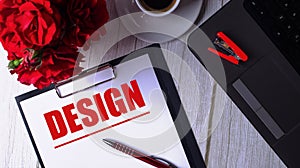 The word DESIGN is written in red on a white notepad near a laptop, coffee, red roses and a pen