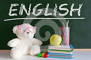 The word `English` written on a blackboard in a language class for early child education. Next, a teddy bear wearing glasses