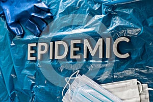 The word epidemic laid by metal letters on crumpled blue pastic with face masks and blue protective glove