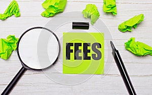 The word FEES written on a green sticky note next to a magnifying glass and a black marker on a wooden table