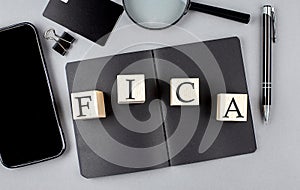 Word FICA on wooden block on black notebook with smartpone, credit card and magnifier