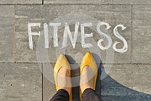 Word fitness written on gray pavement with woman legs, view from above