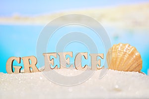 The word Greece on wooden blocks with a seashell in abstract sand with a blurry photo of the sea with mountains. One of the most