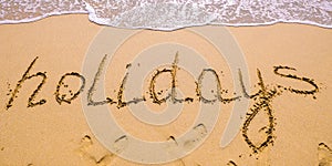 Word Holidays hand written in the sand with a sea wave. Close up sand texture on beach in summer