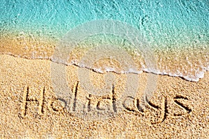 Word HOLIDAYS written on yellow sand, blue sea water wave white foam top view close up, handwriting letters text, summer vacations