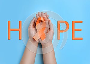 Word HOPE and woman with orange awareness ribbon on light blue background, top view