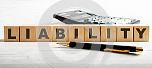 The word LIABILITY is written on the wooden cutouts between the calculator and the pen on a light background